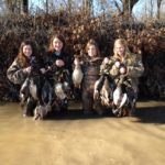 Four female hunters stand in a pond with their harvested ducks.