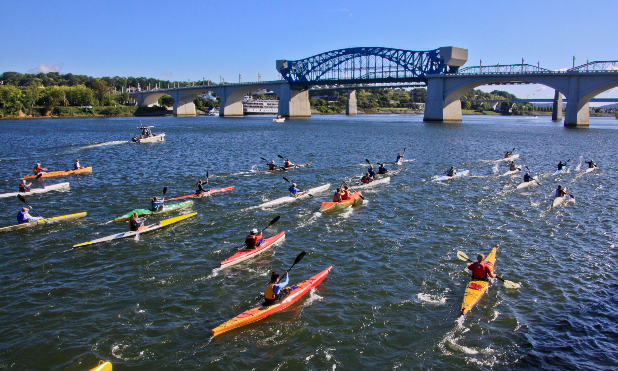 A large group of student kayak on the Tennessee River while a spanning bridge sets the background