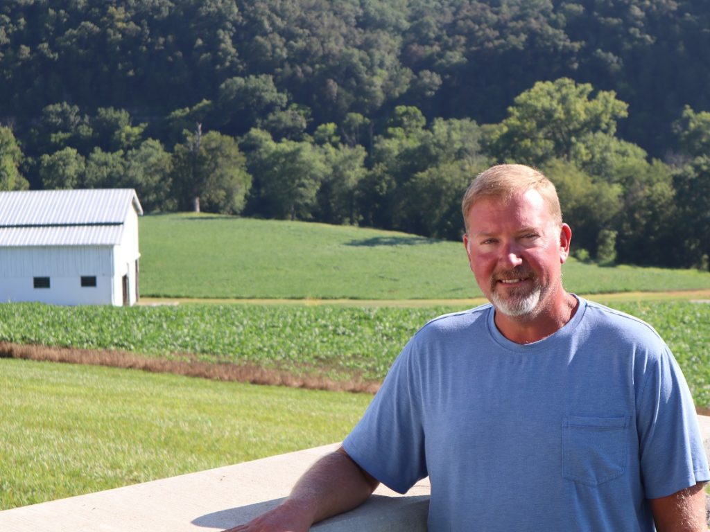 Kyle Owen leans against fence iwth white barn in background on his farm