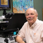 Ernest Bernard sits and smiles at camera with a large microscope on the table behind him.