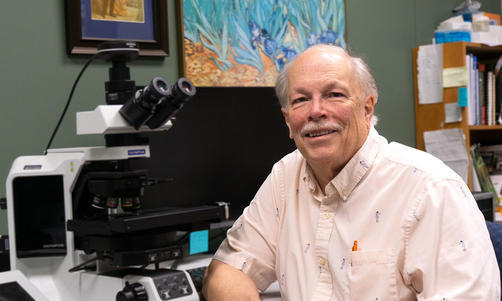 Ernest Bernard sits and smiles at camera with a large microscope on the table behind him.