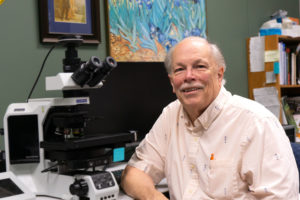 Ernest Bernard sits and smiles at camera with a large microscope on the table behind him.