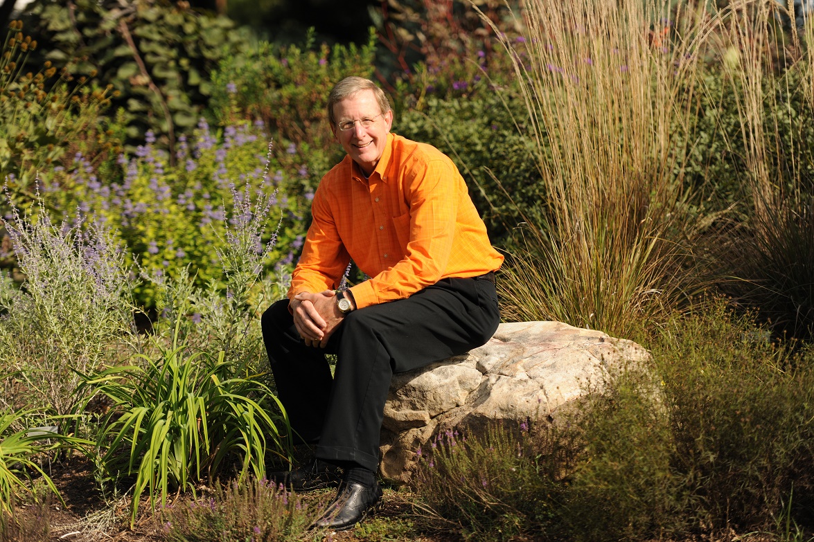 Jim Thompson sits on a rock in a garden, smiling