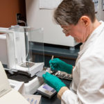 woman in lab wearing white coat and green gloves prepares a soil sample for testing