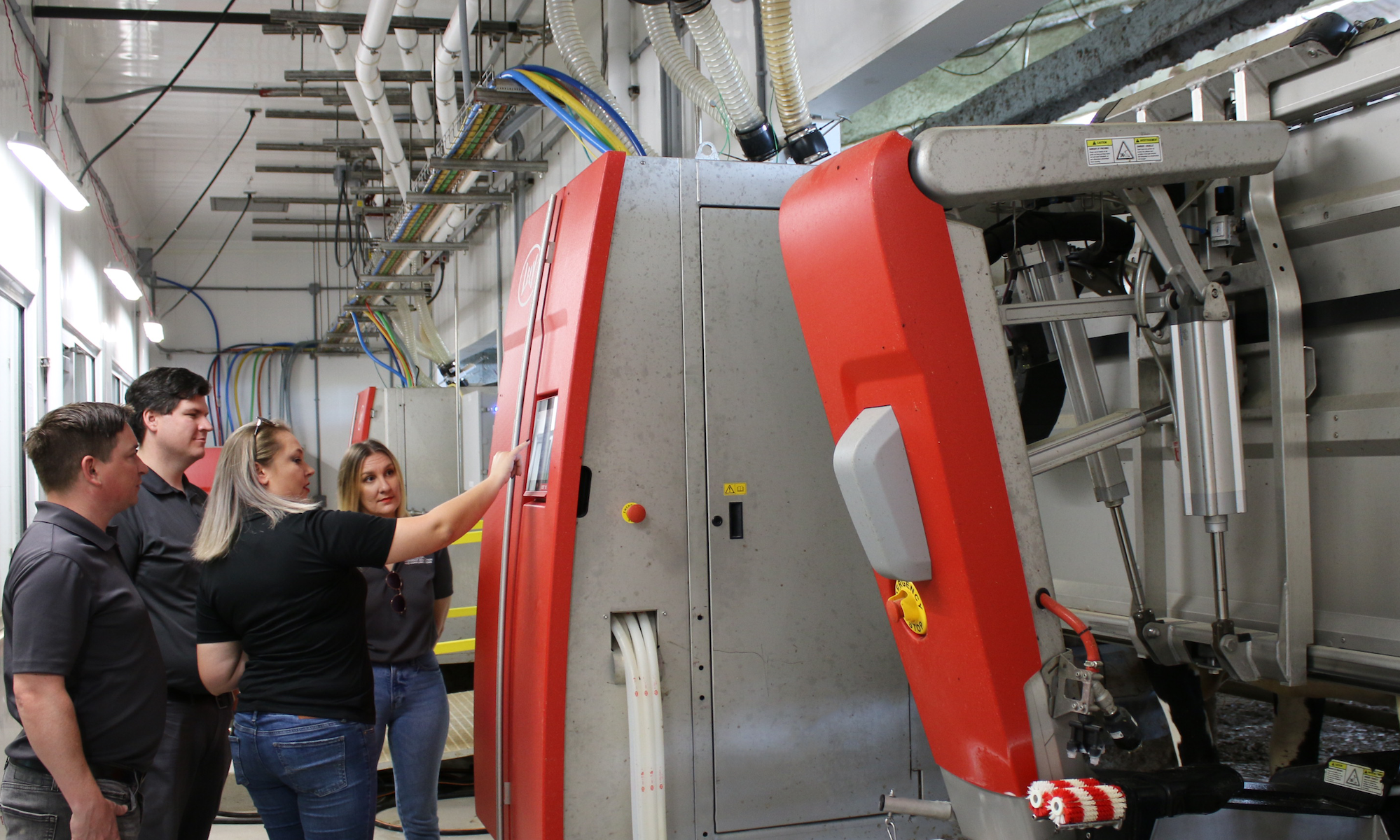 four researchers stand in front of a red and grey robotic milking system. One researcher points to a digital screen on the system