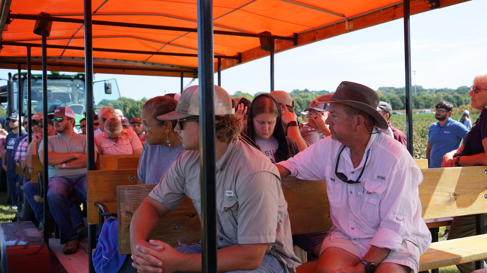 Large group of attendees sit in a wagon while listening to presenters at the 2023 Cotton Tour Field Day.