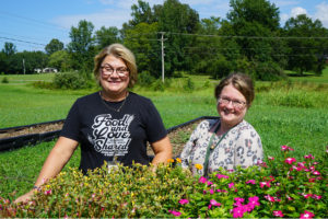 Donna Vick (left) and Jennifer Thornton (right) stand behind a raised flower bed with yellow and pink flowers. trees and powerlines in the background