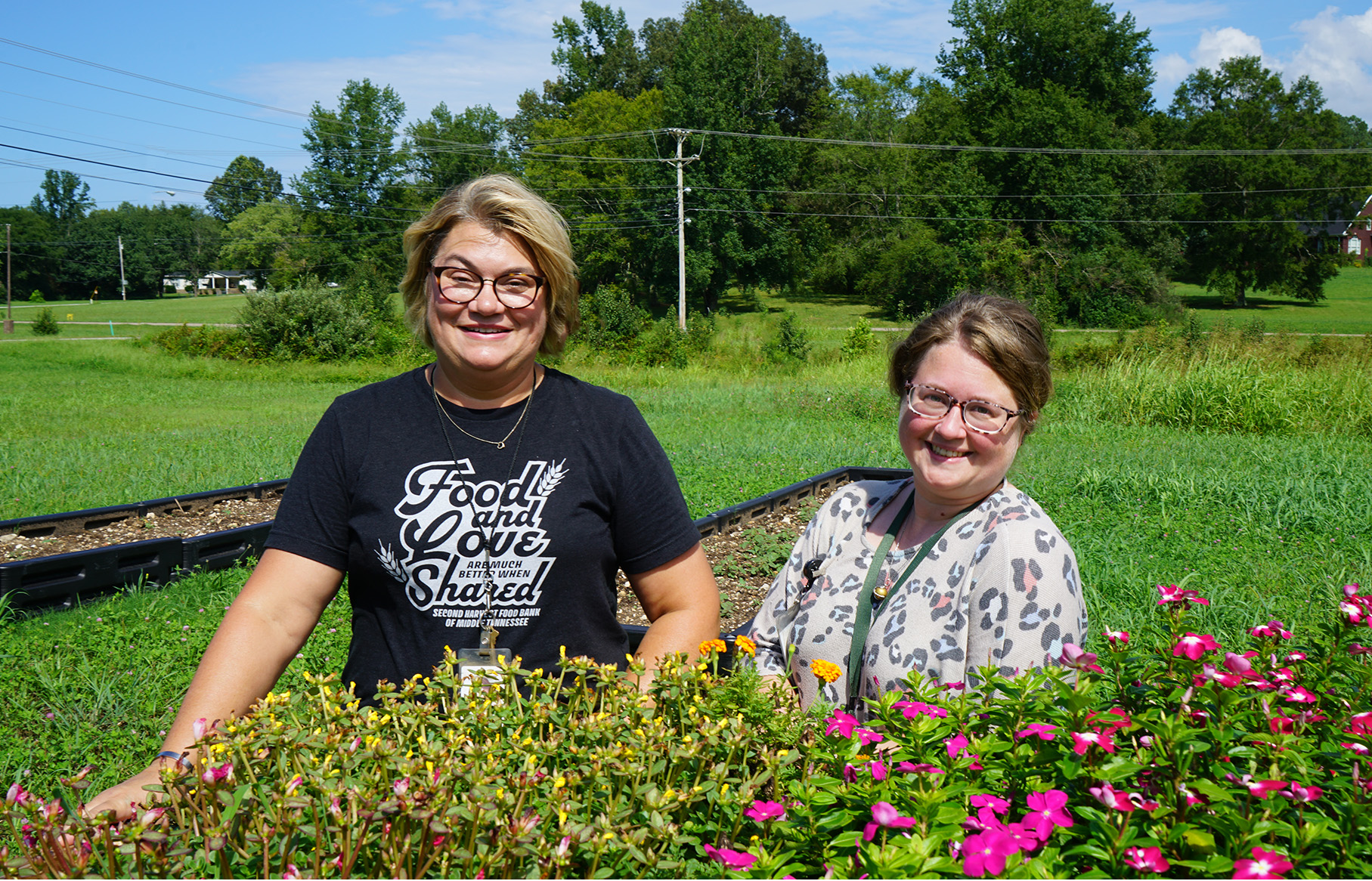 Donna Vick (left) and Jennifer Thornton (right) stand behind a raised flower bed with yellow and pink flowers. trees and powerlines in the background