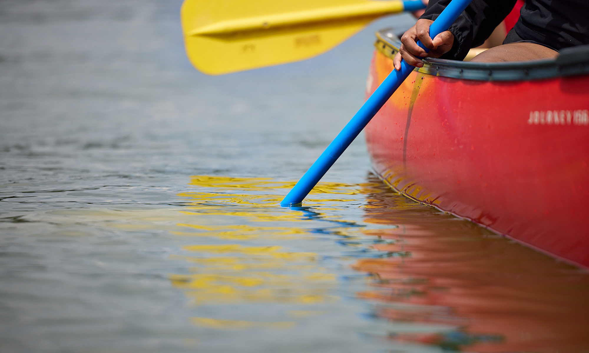 Side hull of orange canoe with blue and yellow paddles dipping into the water