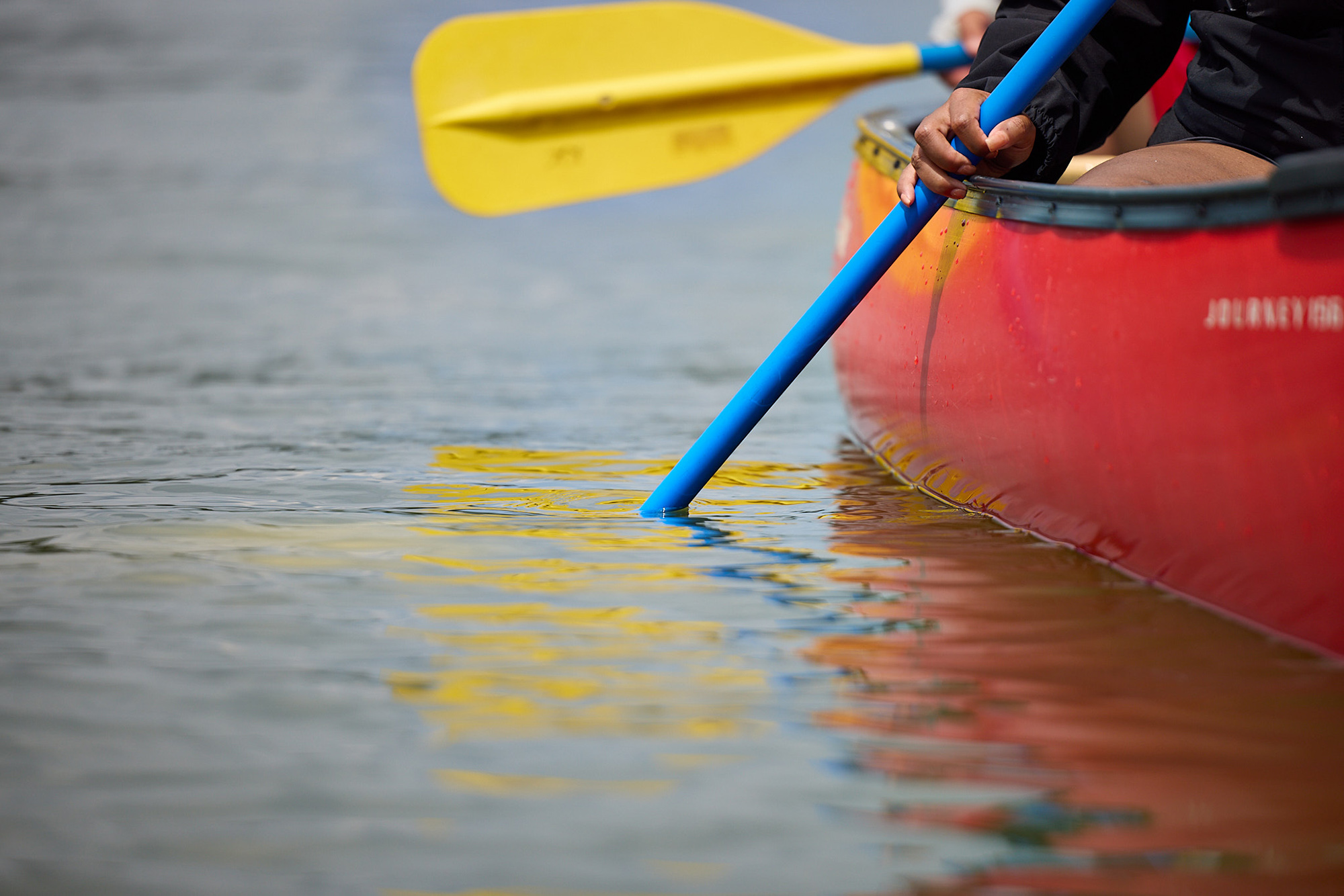 Side hull of orange canoe with blue and yellow paddles dipping into the water