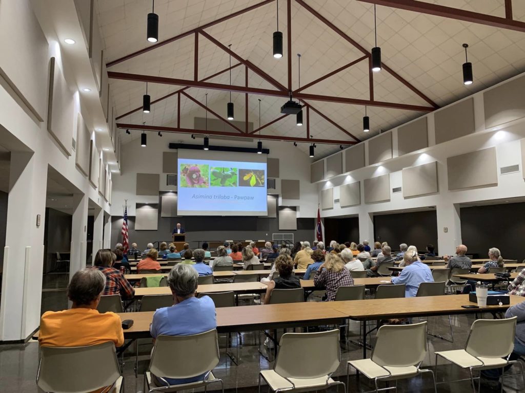 Inside a large lecture hall with high ceilings, people sit at rows of tables facing a person at the front of the room who is standing at a lectern. A screen behind the presenter has images of flowers and fruits projected on it with the text "Asimina triloba - Pawpaw" below