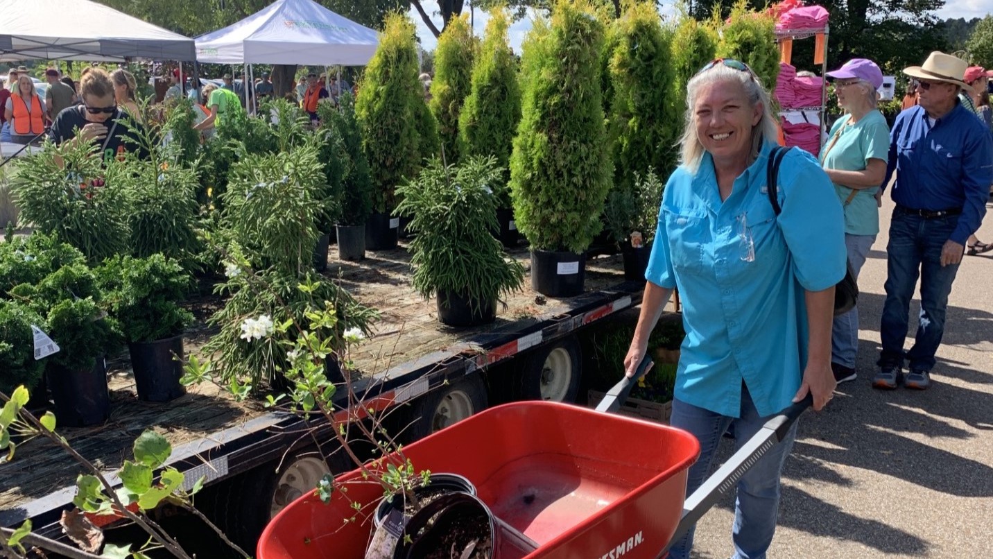 Smiling woman in blue shirt pushes a red wheelbarrow full of plants. Rows of potted trees and assorted plants are behind her.