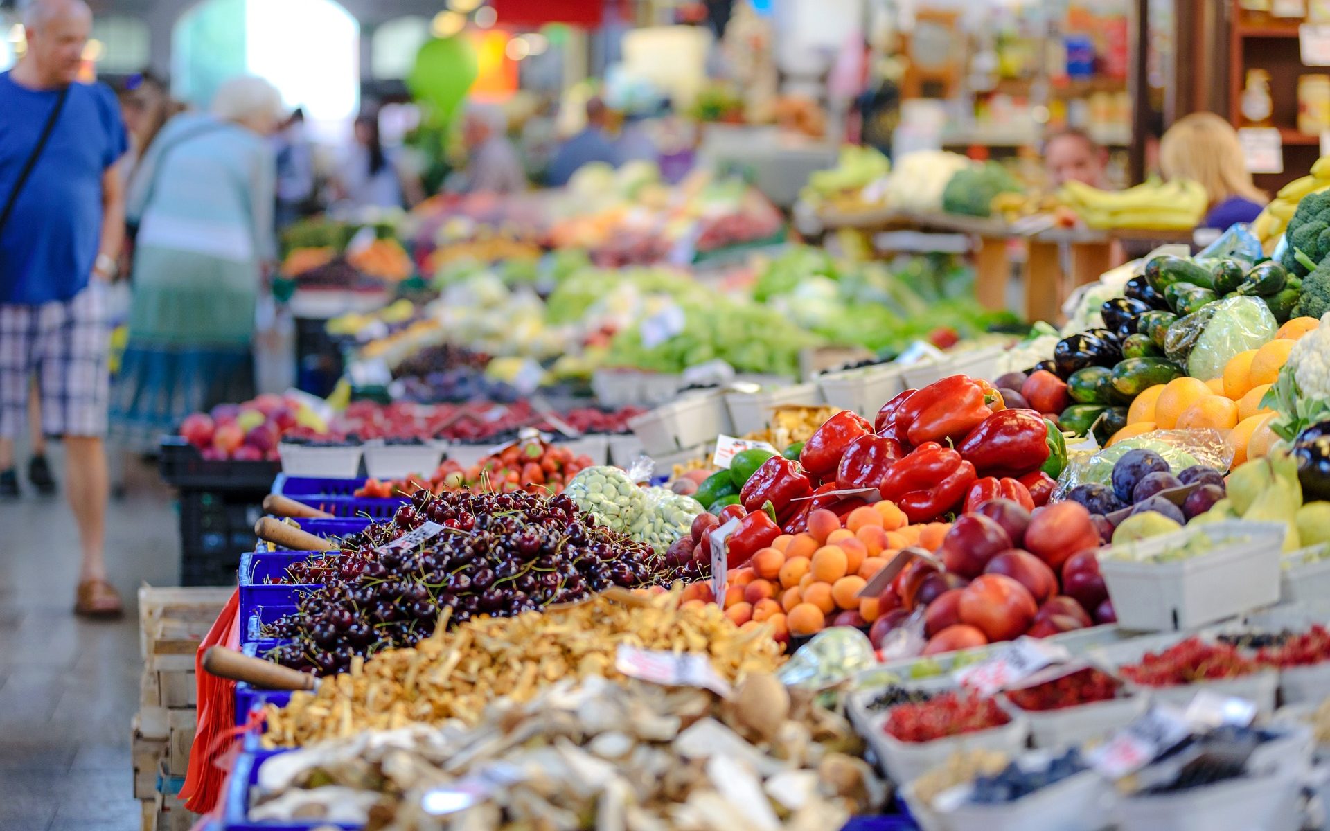 produce section at a market showing berries, apricots, cucumbers and peppers with shoppers in the background