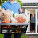 person holding a basket of groceries stands outside a house where a person opens the door