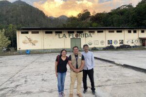 María Franco Escobar, Edwin Villa, and Carlos Trejo-Pech stand in front of a sunset and a wall that has a mural reading 