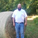 A man in a white shirt stands in front of a hay bale