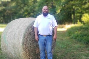 A man in a white shirt stands in front of a hay bale