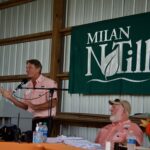 A speaker stands at a podium during Milan No-Till Field Day, a green banner that says "Milan No-Till" is positioned on the wall behind him