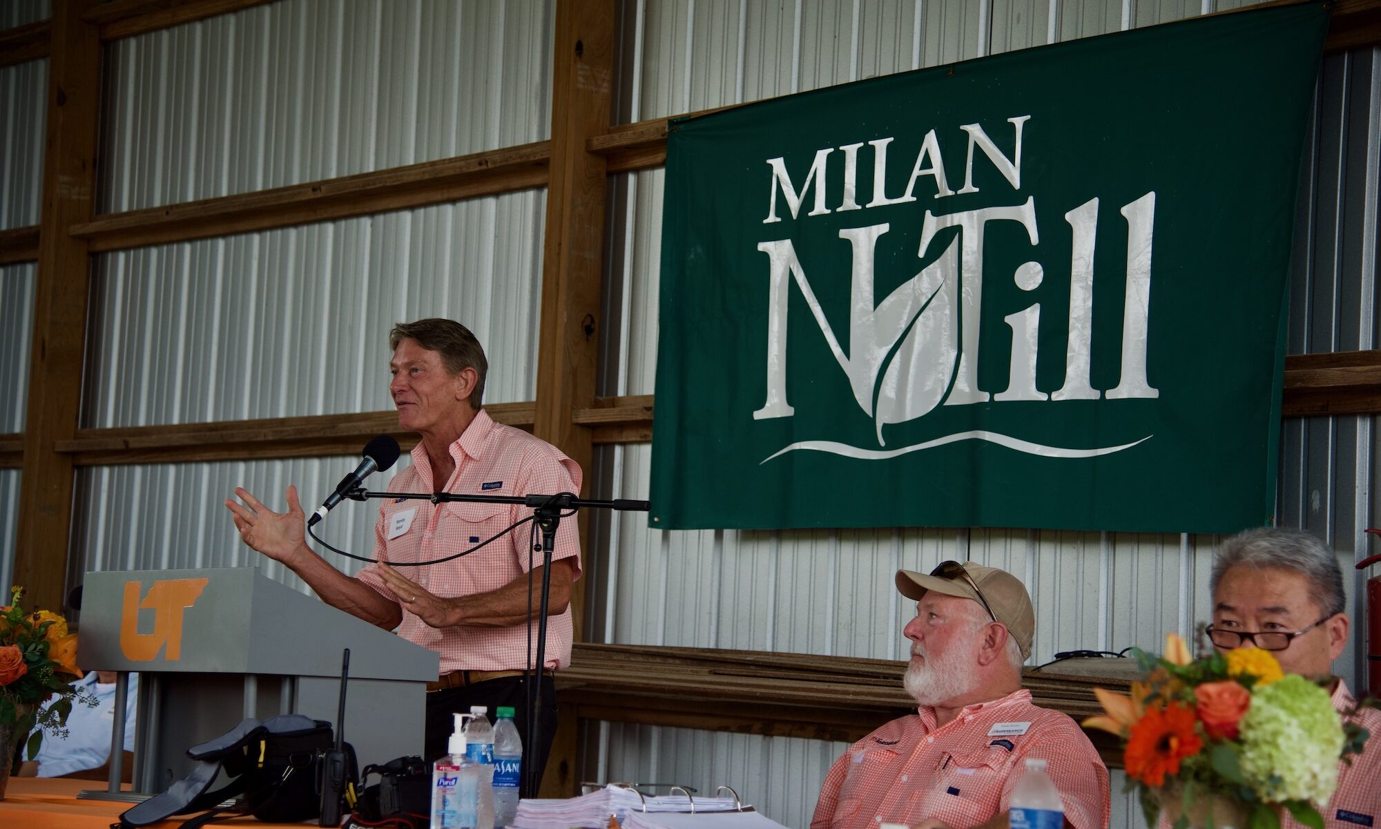 A speaker stands at a podium during Milan No-Till Field Day, a green banner that says "Milan No-Till" is positioned on the wall behind him