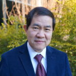 A man in a suit poses in front of a yellow bush outside