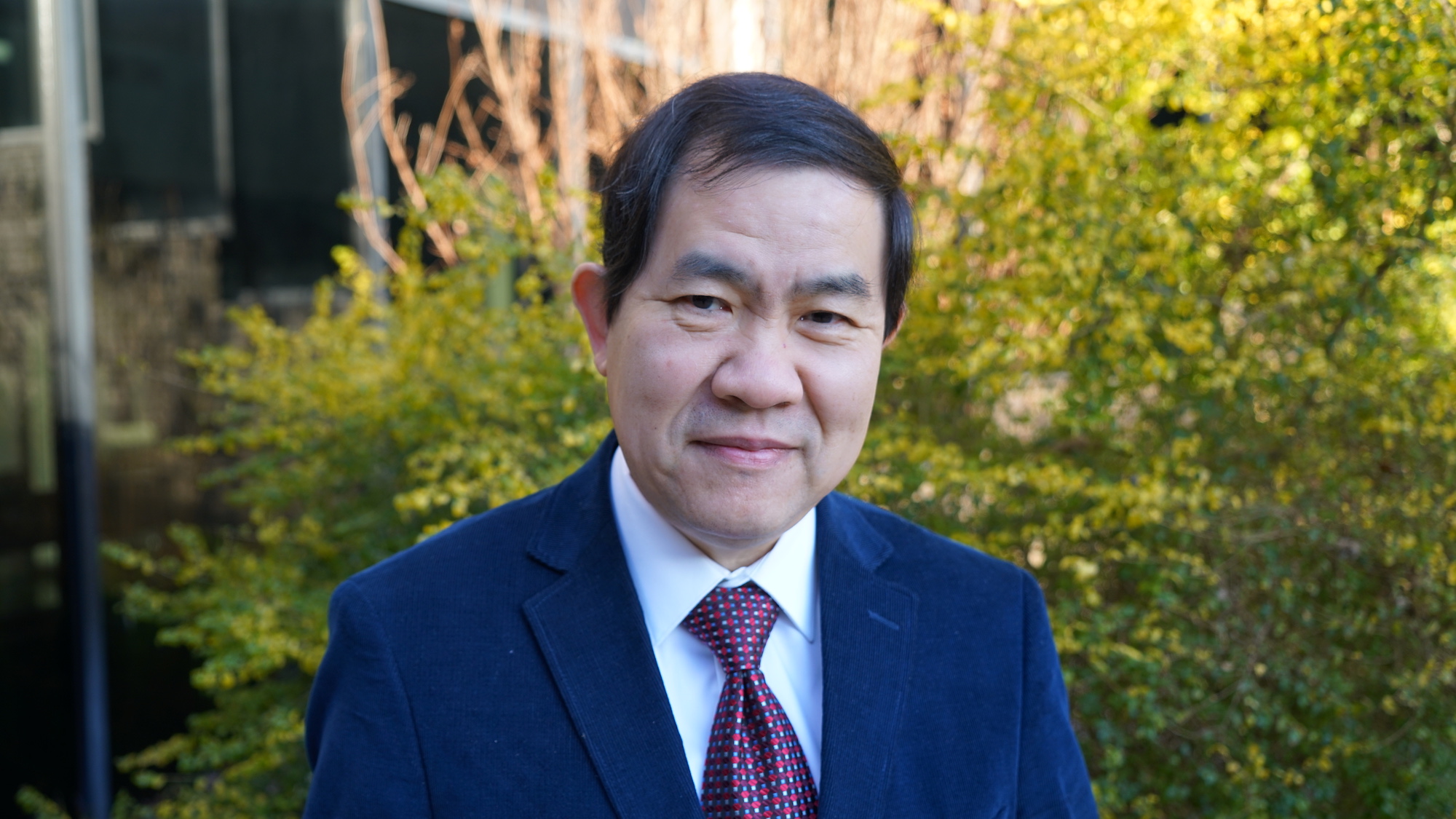 A man in a suit poses in front of a yellow bush outside