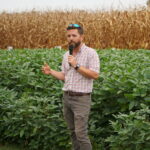 A man with a microphone presents in front of a cotton and corn field