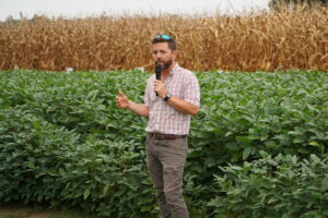 A man with a microphone presents in front of a cotton and corn field