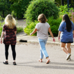 Three women with their backs turned walk down a road next to a garden