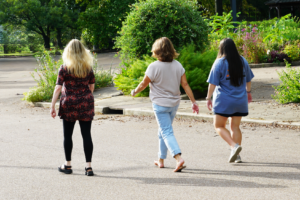 Three women with their backs turned walk down a road next to a garden