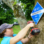 Woman with an electric drill installs a mile marker sign onto a tree