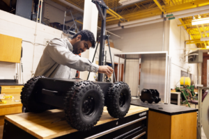 A man works on a black, four-wheeled robot on a table inside his research lab