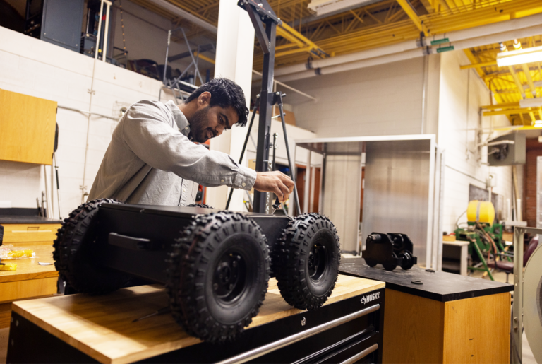 A man works on a black, four-wheeled robot on a table inside his research lab