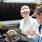 Two researchers, one holding a clipboard, examine a plant in a greenhouse