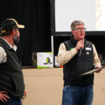 Two presenters, one with a microphone, speak in front of a crowd