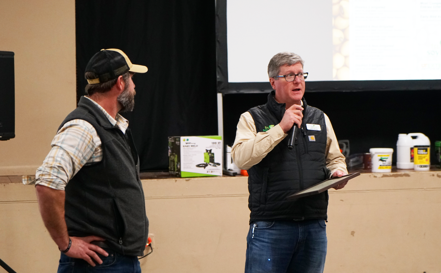 Two presenters, one with a microphone, speak in front of a crowd