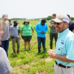 A researcher in a blue shirt holds a small plant as he teaches amongst a circle of college students
