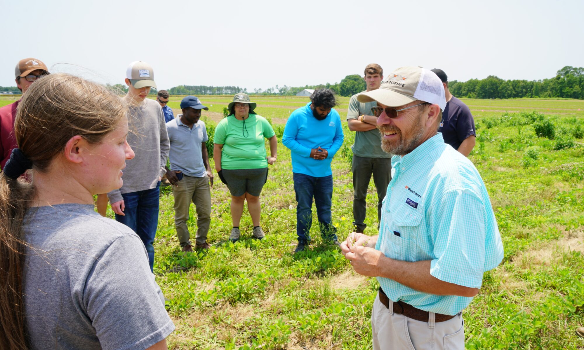 A researcher in a blue shirt holds a small plant as he teaches amongst a circle of college students