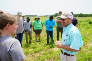 A researcher in a blue shirt holds a small plant as he teaches amongst a circle of college students