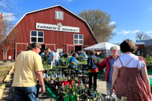 A crowd shops plants outside a red barn with a sign reading 