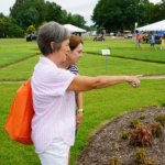 Two women point at plants at UT Gardens, Jackson, with white Summer Celebration tents in the background