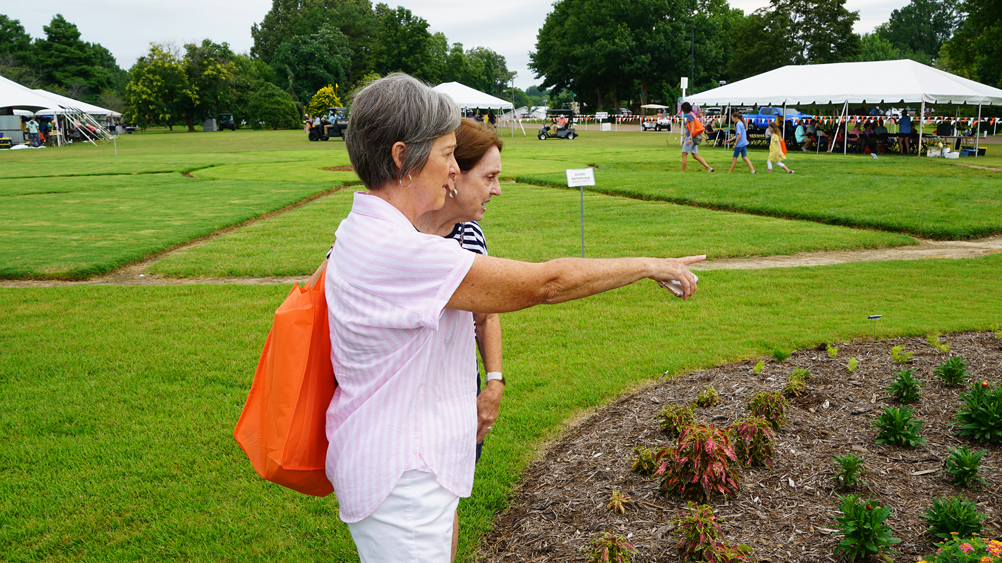 Two women point at plants at UT Gardens, Jackson, with white Summer Celebration tents in the background