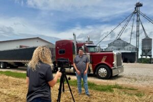rachel painter taking a video of Aaron Smith in front of a semi truck and grain bins