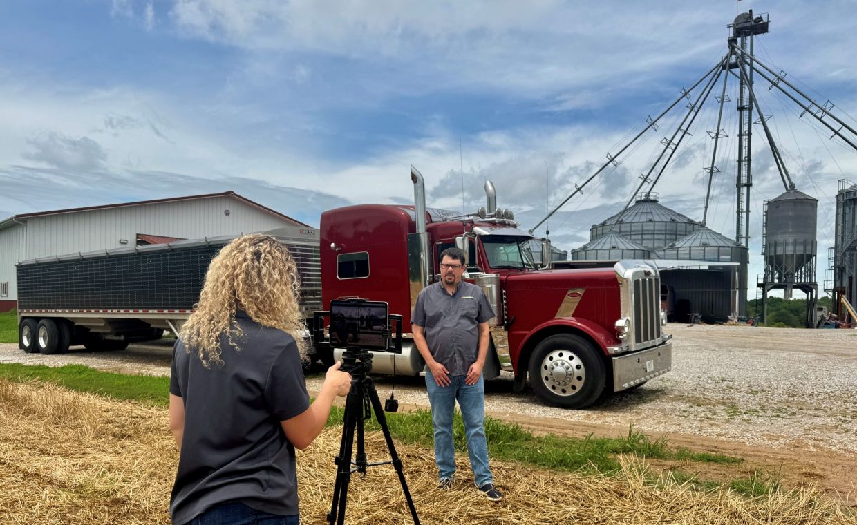 rachel painter taking a video of Aaron Smith in front of a semi truck and grain bins