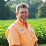 A man in an orange shirt poses for a professional headshot in front of a green cotton field