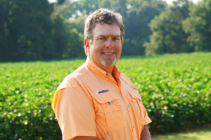 A man in an orange shirt poses for a professional headshot in front of a green cotton field