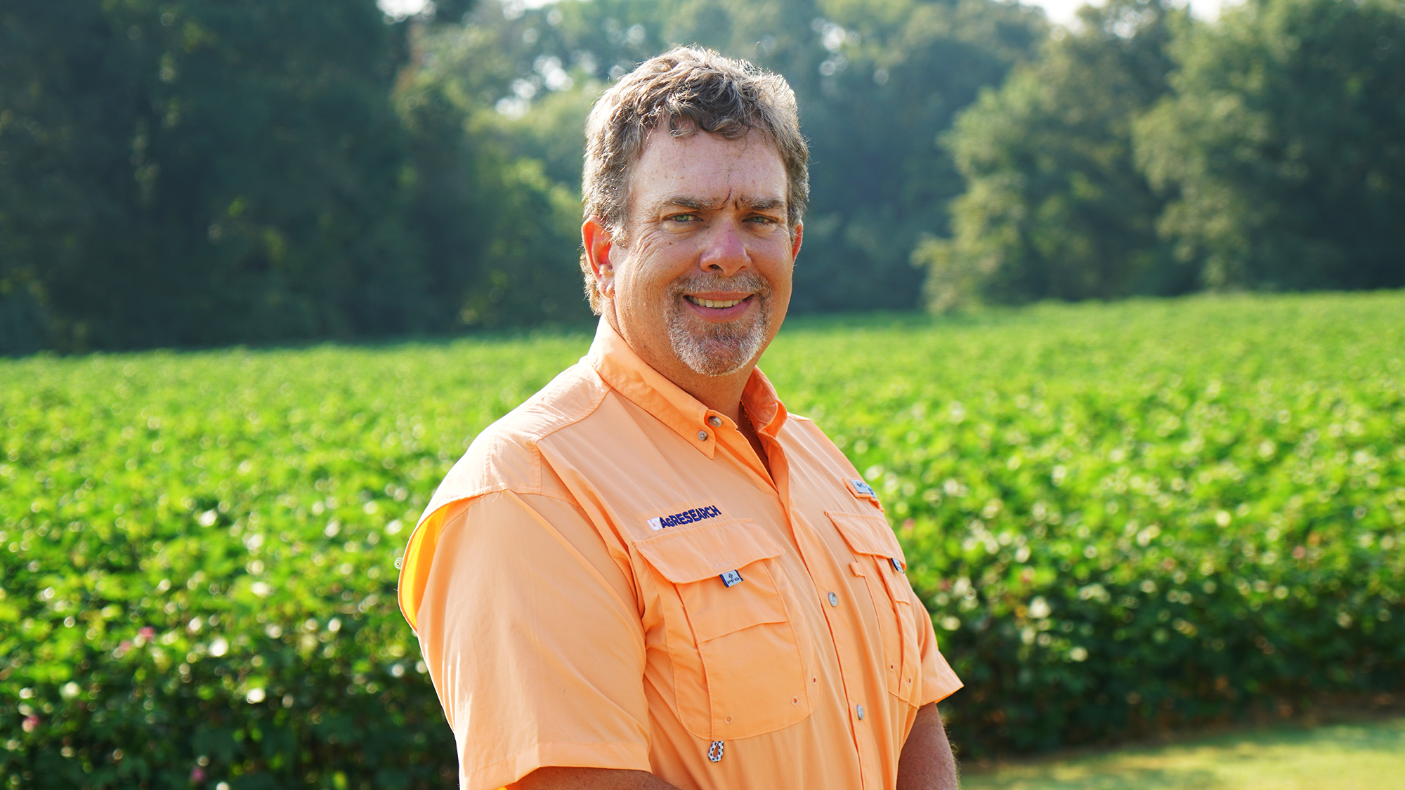 A man in an orange shirt poses for a professional headshot in front of a green cotton field