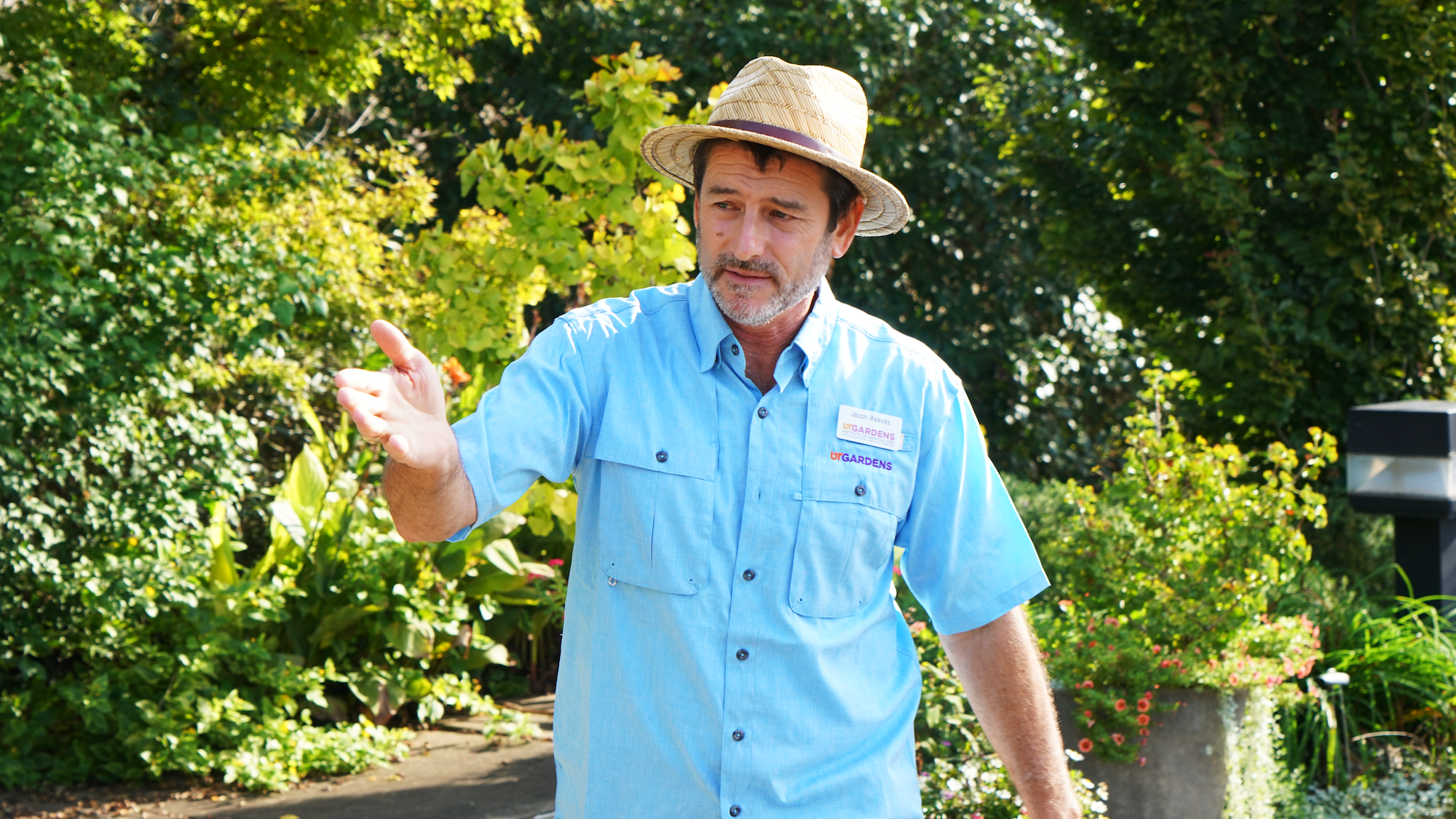 A man in a hat and blue shirt gestures at a collection of plants while giving an outdoor garden tour