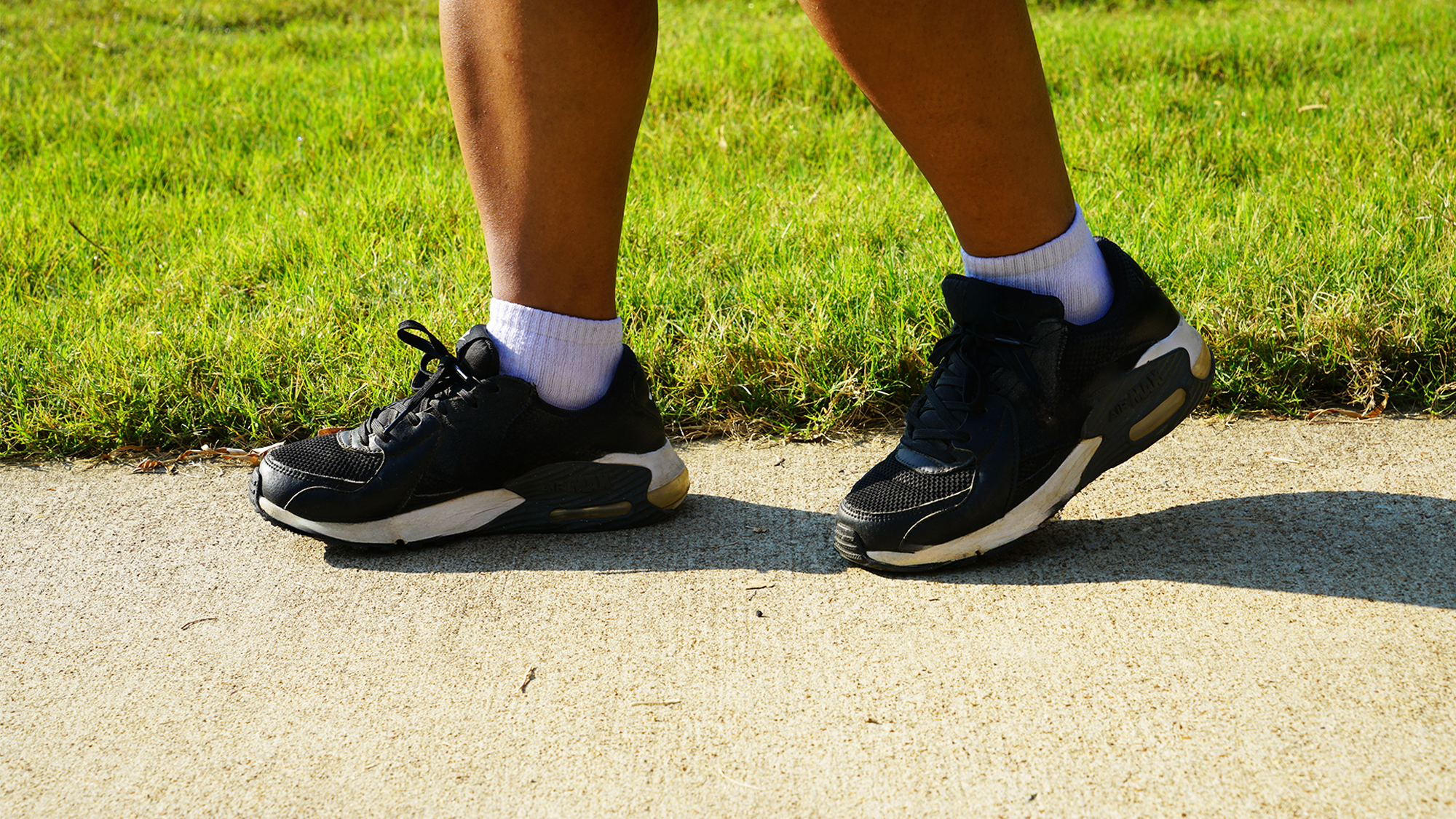 A close up of a person's black and white shoes as they walk on a concrete path next to a green lawn