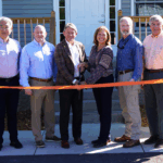 Eight people, two holding a pair of scissors, pose for a picture behind an orange ribbon at the front of a house