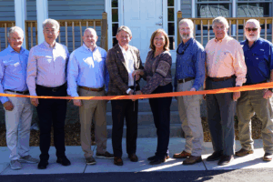 Eight people, two holding a pair of scissors, pose for a picture behind an orange ribbon at the front of a house